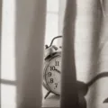 Jam weker di atas meja samping tempat tidur yang tersiram cahaya matahari pagi untuk ilustrasi cara mengatur jadwal tidur