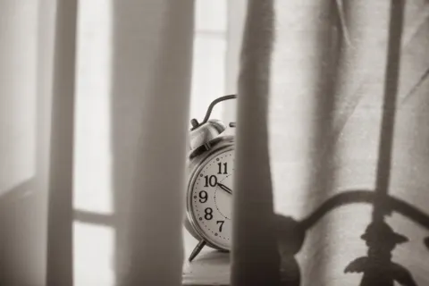 Jam weker di atas meja samping tempat tidur yang tersiram cahaya matahari pagi untuk ilustrasi cara mengatur jadwal tidur