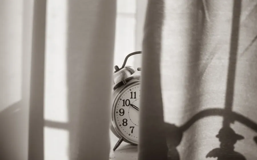 Jam weker di atas meja samping tempat tidur yang tersiram cahaya matahari pagi untuk ilustrasi cara mengatur jadwal tidur
